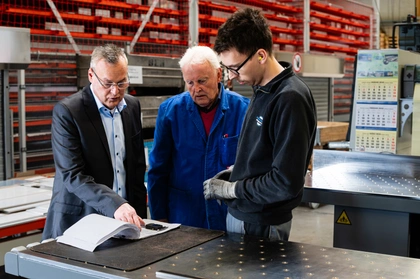 Drei Mitarbeiter der Aluminium Halbzeuge und Zuschnitte GmbH besprechen an der Werkbank einen Auftrag, im Hintergrund die Lagerregale mit Zuschnitten