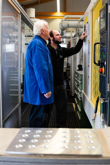 Zwei Mitarbeiter an der Steuerung einer CNC-Bearbeitungsmaschine, der ältere erklärt dem jüngeren die Eingabe