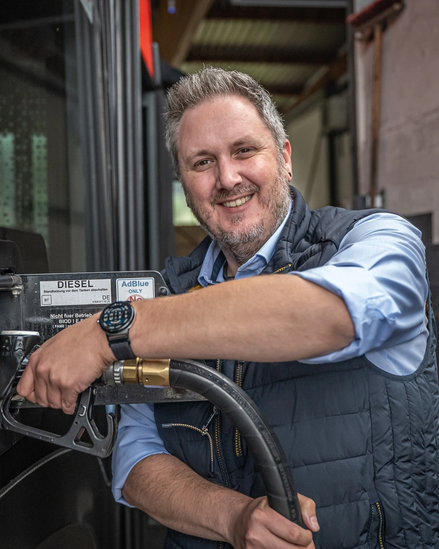 Mann in Weste lehnt lächelnd an der Dieselzapfsäule im Busdepot
