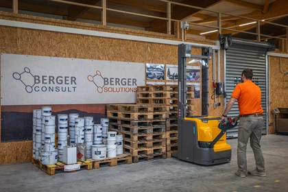 Ein Mitarbeiter von Berger-Consult zieht einen Elektro-Hubwagen durch das Materiallager, an der Wand das Firmenbanner, davor gestapelte Paletten und Gebinde