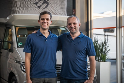 Zwei Mitarbeiter im Verkaufsraum eines Caravanhändlers vor einem Wohnmobil, beide in Poloshirts mit Firmenlogo