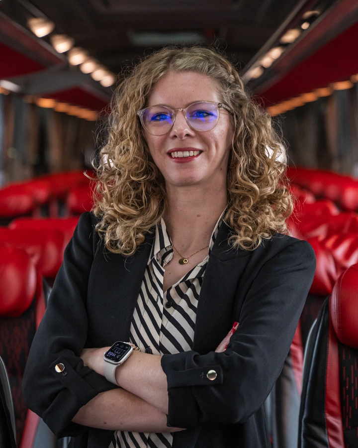 Frau mit Brille und verschränkten Armen steht lächelnd im Gang eines Reisebusses