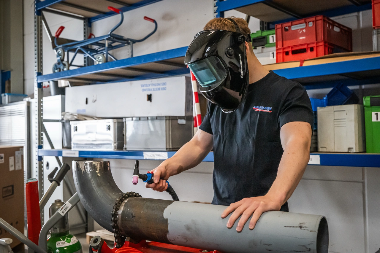 Ein junger Schweißer mit Schweißhelm schweißt in der Werkstatt einen Rohrbogen