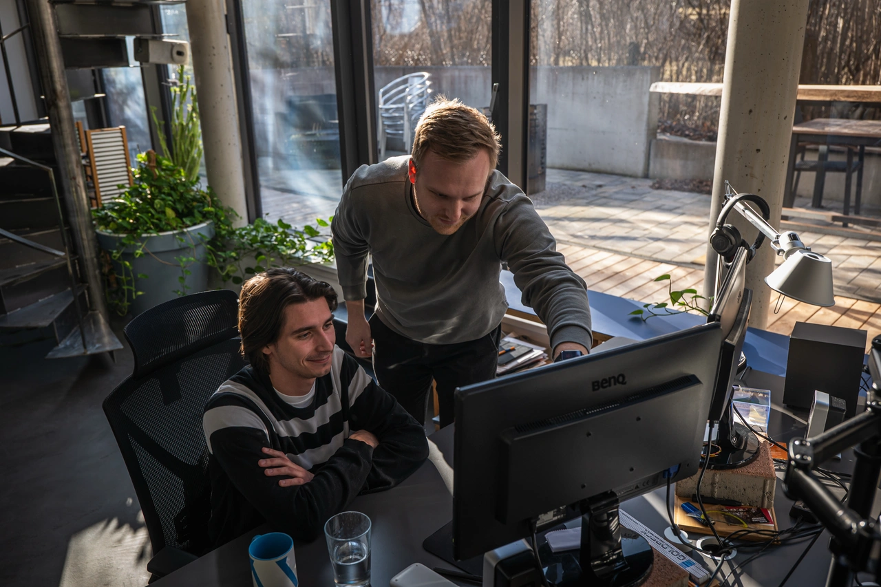 Zwei Kollegen gehen im Büro gemeinsam etwas am Bildschirm durch, einer zeigt auf den Monitor