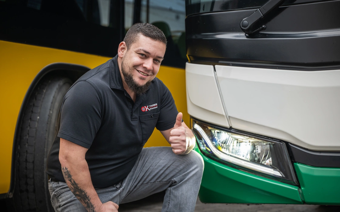 Mitarbeiter von O.K.-Reisen Kleinhenz kniet vor einem Linienbus und hebt den Daumen