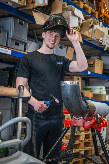 Junger Schweißer in schwarzem T-Shirt mit hochgeklapptem Schweißhelm hält einen Brenner, vor ihm ein eingespanntes Stahlrohr, dahinter Regale mit Material