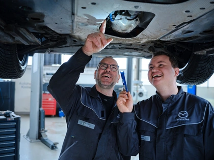 Ein erfahrener Kfz-Mechaniker mit Brille zeigt einem jungen Kollegen lachend etwas am Unterboden eines Autos auf der Hebebühne, der Jüngere hält eine Stablampe