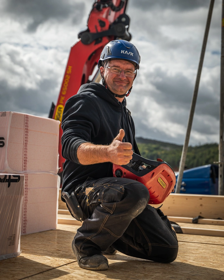 Handwerker mit Helm kniet auf einer Holzdecke, hält eine Kranfernsteuerung und zeigt den Daumen nach oben