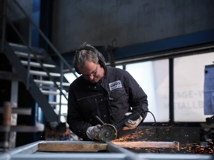 Ein Metallbauer mit Gehörschutz und Schutzbrille trennt mit dem Winkelschleifer an einem Stahlrahmen, die Funken fliegen über die Werkbank, dahinter die Montagehalle