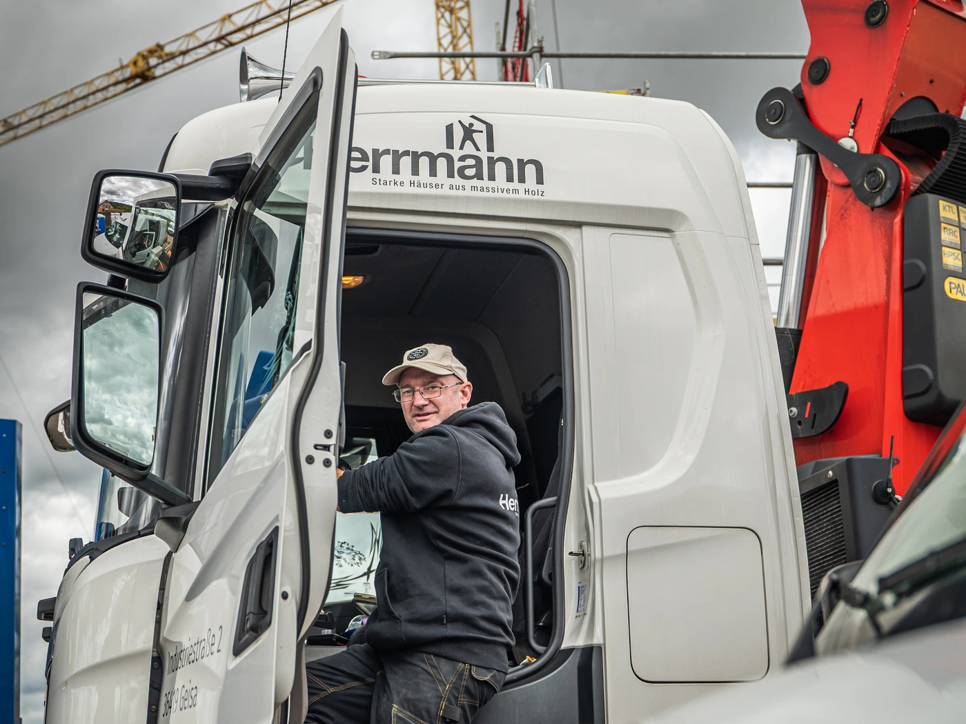 Mann mit Schirmmütze steigt in das Fahrerhaus eines Lkw von Herrmann Holzbau, rechts der rote Ladekran