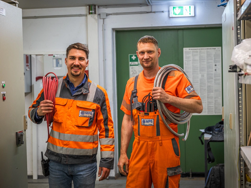 Zwei Kollegen in Arbeitskleidung tragen Kabelrollen durch einen Technikraum und lächeln in die Kamera