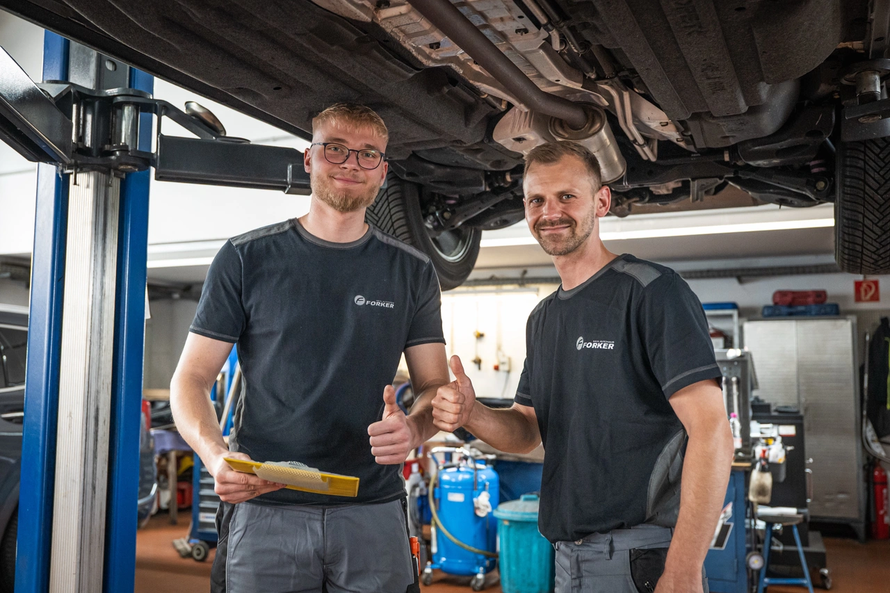Zwei Mechaniker unter einer Hebebühne, einer hält eine gelbe Mappe, beide zeigen den Daumen nach oben