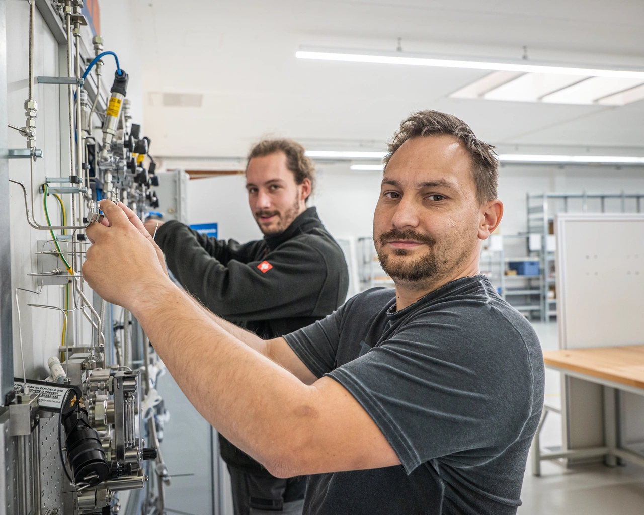 Techniker justiert Ventile an einer Messtafel, ein Kollege arbeitet dahinter mit