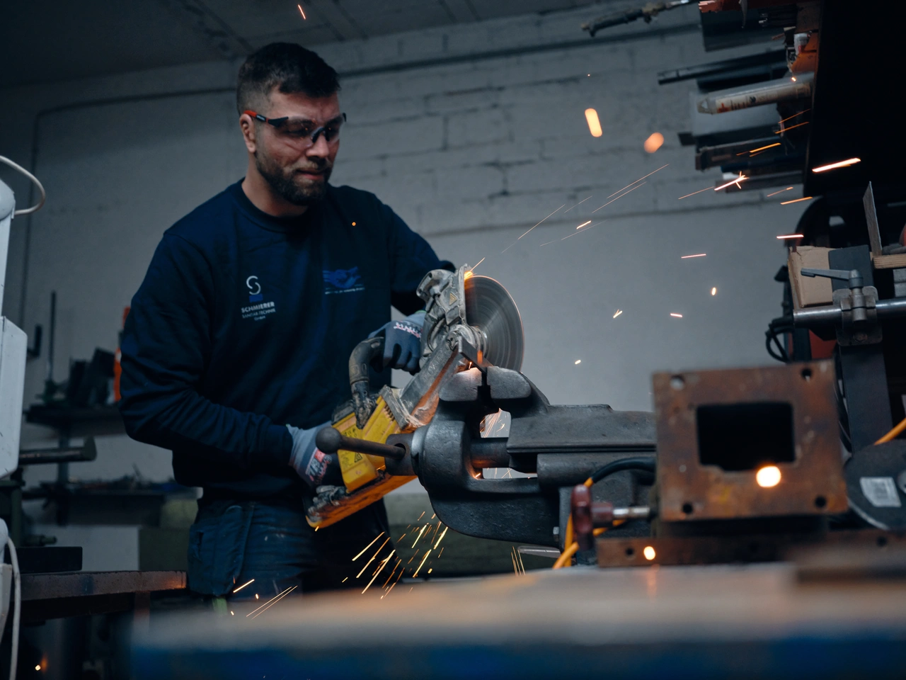 Ein Metallbauer mit Schutzbrille trennt ein Werkstück am Schraubstock, Funken fliegen