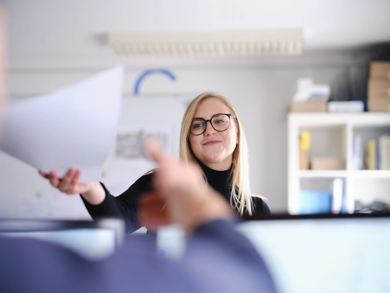 Eine Mitarbeiterin im Büro reicht ein Blatt Papier über den Schreibtisch