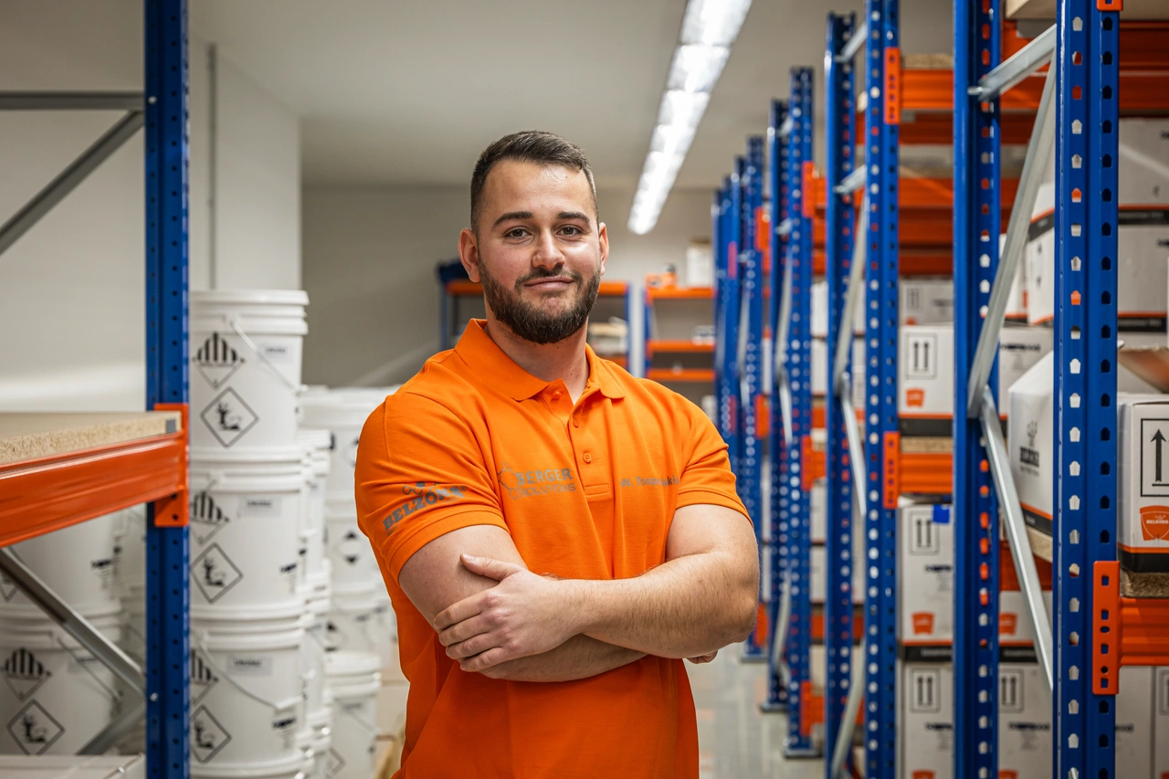Ein Techniker in orangefarbenem Poloshirt steht mit verschränkten Armen zwischen Regalen voller Farbeimer im Lager