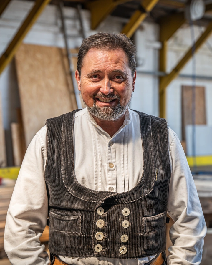 Lächelnder Zimmermann mit grauem Bart in Cordweste und weißem Hemd steht in der Werkhalle vor Holzplatten