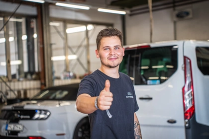 Ein junger Kfz-Mechaniker in dunklem Poloshirt mit Firmenlogo zeigt in der Werkstatthalle vom Autohaus MOSIG den Daumen nach oben, hinter ihm ein weißer Ford-Transporter