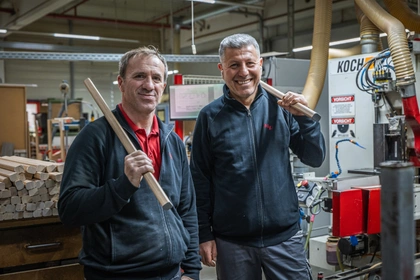 Zwei Mitarbeiter in der Holzbearbeitung halten fertig profilierte Rundstäbe in den Händen, dahinter eine Profiliermaschine