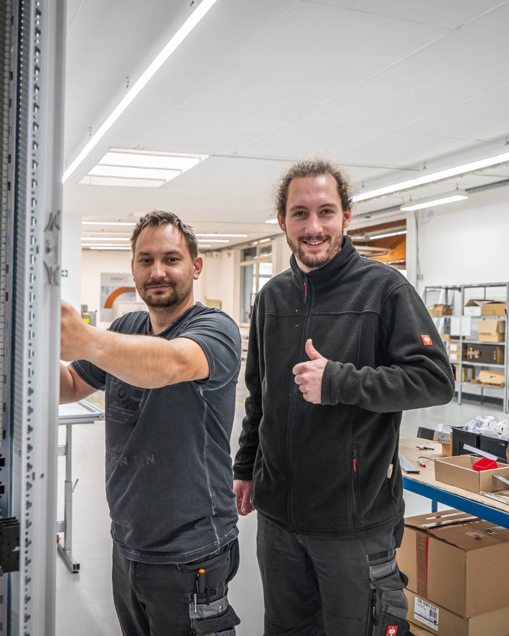 Zwei Techniker an einem Schaltschrank in einer hellen Werkhalle, einer hebt den Daumen
