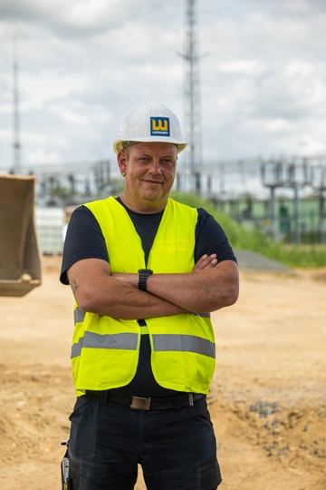 Bauarbeiter mit weißem Schutzhelm und gelber Warnweste steht mit verschränkten Armen auf einer Baustelle, im Hintergrund ein Umspannwerk