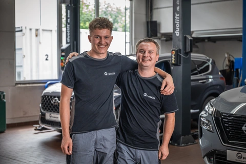 Zwei junge Kfz-Mechaniker in dunklen Arbeitsshirts stehen lachend Arm in Arm in einer Werkstatt vor Hebebühnen